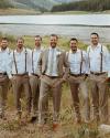 Six men in dress shirts and khaki pants, one in a suit, stand together outdoors by a lake with mountains and trees in the background.