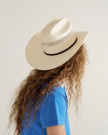 Person with long, curly hair wearing a light-colored cowboy hat and a blue T-shirt, facing away from the camera against a plain background.