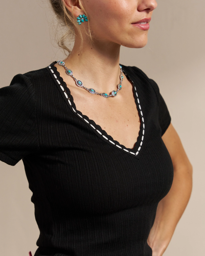 A woman in a black V-neck shirt, blue flower earrings, and a blue bead necklace is posing with her hands on her hips.