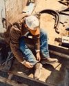 A person wearing a brown jacket, blue jeans, and work boots sits outdoors on metal steps, adjusting a boot. Tools and hoses are scattered nearby on the ground.