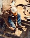 A person wearing a brown jacket, blue jeans, and work boots sits outdoors on metal steps, adjusting a boot. Tools and hoses are scattered nearby on the ground.