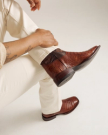 The William in Dark Pecan Caiman. Person wearing cream pants and The William Caiman brown crocodile-patterned leather boots, sitting cross-legged with a tattoo visible on their forearm.
