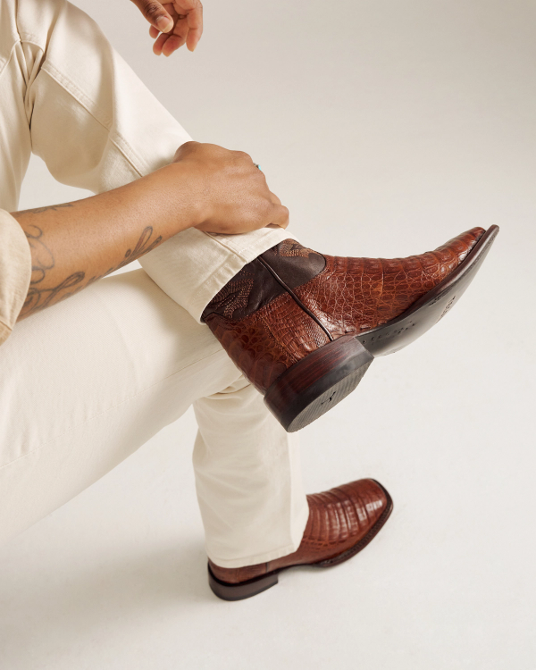 The William in Dark Pecan Caiman. Person wearing cream pants and The William Caiman brown crocodile-patterned leather boots, sitting cross-legged with a tattoo visible on their forearm.