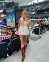 A woman in a white dress and red cowboy boots stands by stadium seats. Concert stage in the background displays "Kenny Chesney" with people seated and standing nearby.