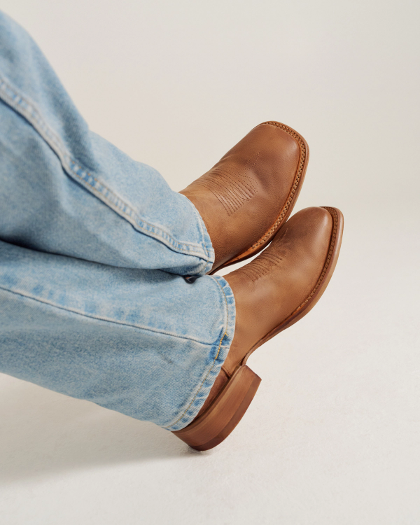 Person wearing light blue jeans and brown leather boots with a low heel, feet crossed and resting against a plain light background.