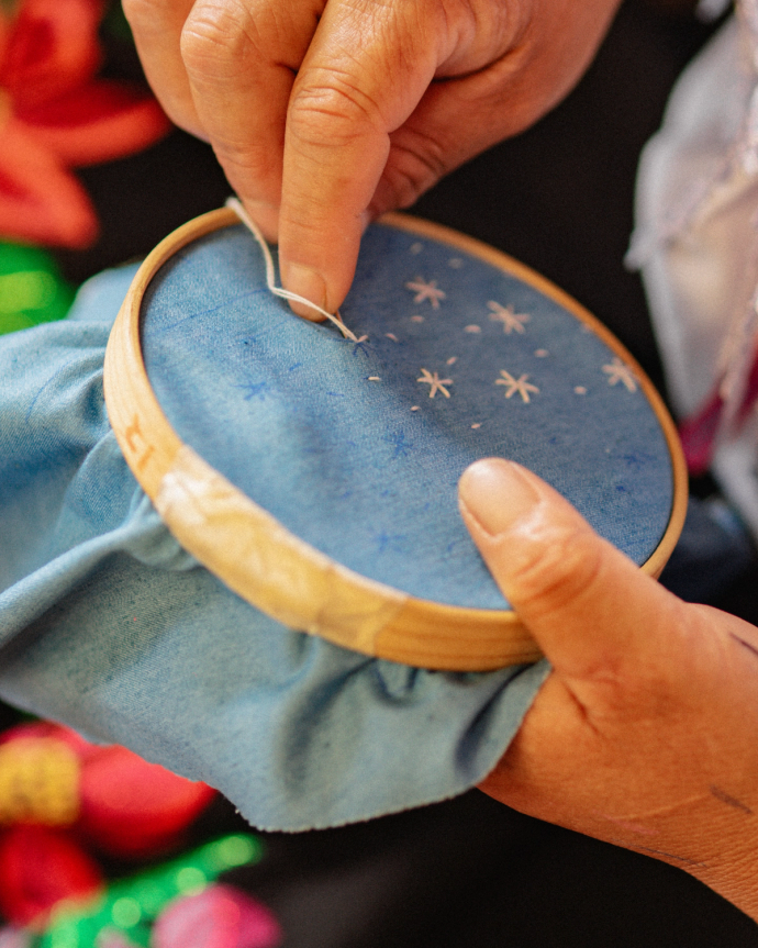 A person hand-embroiders small white star shapes on blue fabric stretched in an embroidery hoop.