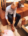 A man sitting on a brown leather couch tries on a pair of brown cowboy boots, with an opened boot box on a table nearby.