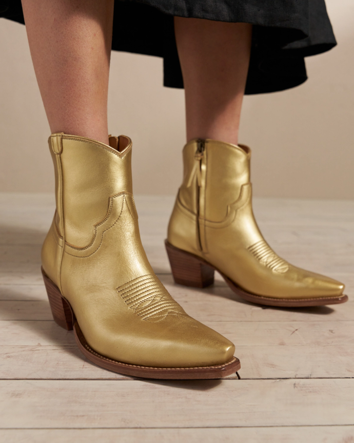 Person wearing gold cowboy boots with wooden heels on a light wooden floor.