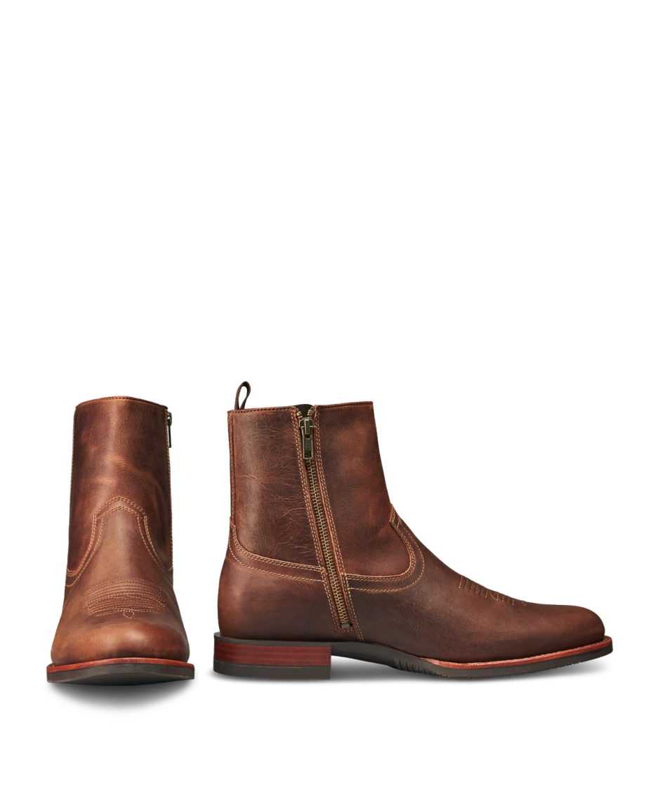 The Wayne in Maplewood Cowhide. Pair of brown cowhide leather ankle boots with side zippers, worn finish, round toes, and low maplewood heels. One boot is shown from the front, the other from the side.
