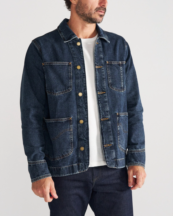 A man wearing a dark denim chore jacket over a white t-shirt and dark jeans stands against a plain background.