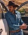 A person in a cowboy hat and denim shirt leans against a truck door outdoors on a sunny day.