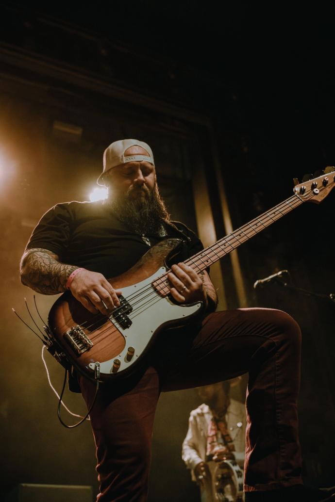 A bearded man playing an electric bass guitar on stage, intensely focused, with stage lighting highlighting him from behind.