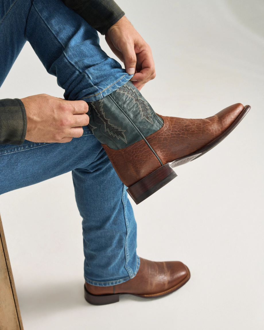 The Doc in Burnt Umber / Blue Cowhide. A person wearing blue jeans adjusts a Burnt Umber / Blue Cowhide leather cowboy boot while seated on a wooden stool.