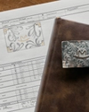 A detailed metal belt buckle, a brown leather-bound book, and a printed order form with a sketched buckle design rest on a wooden surface.