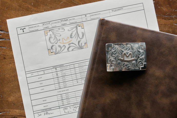 A detailed metal belt buckle, a brown leather-bound book, and a printed order form with a sketched buckle design rest on a wooden surface.