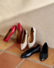 Three pairs of pointed-toe flats in red, white, and black are arranged against a light-colored textured wall on a tiled floor.