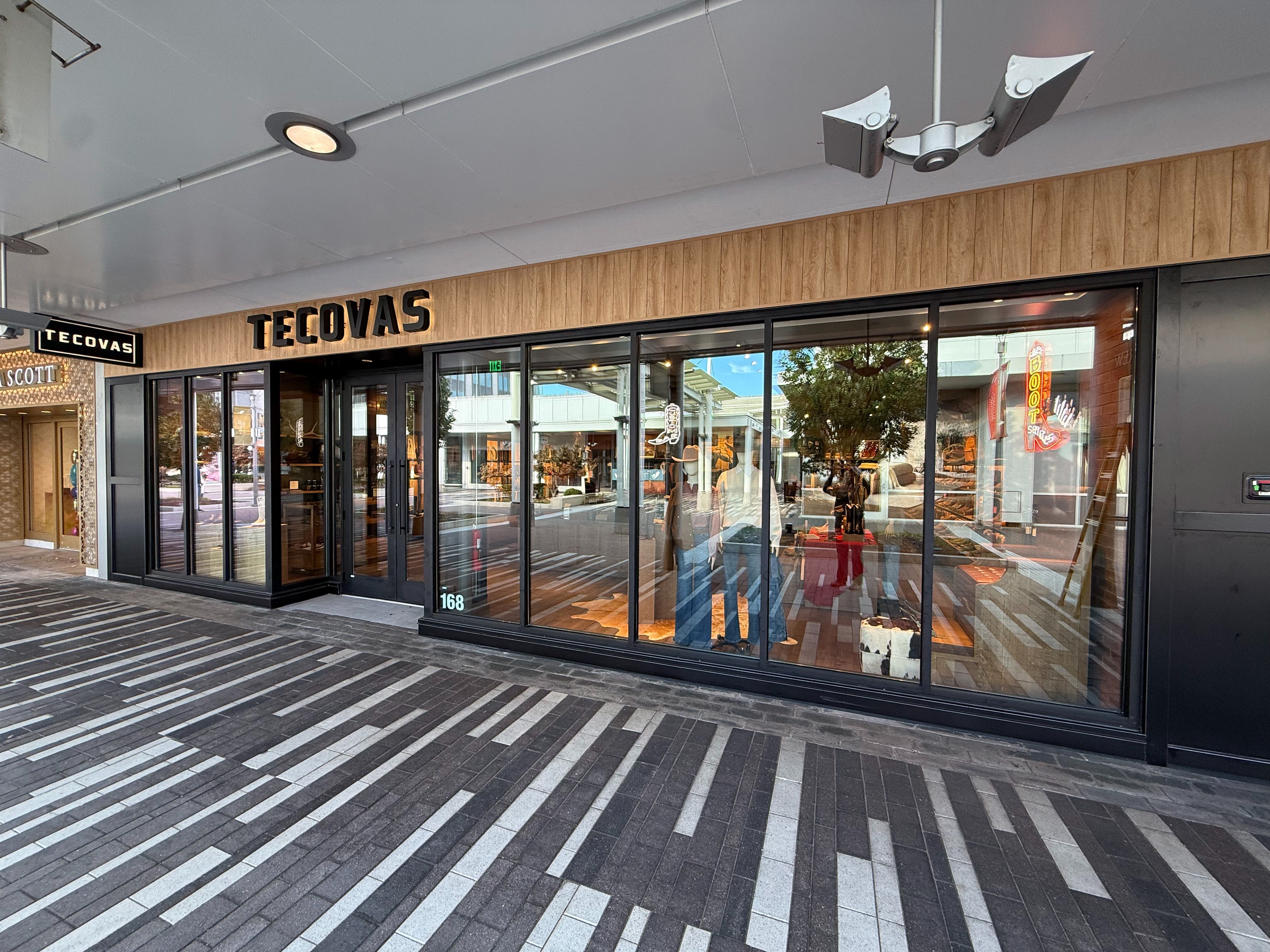 Storefront of Tecovas with large glass windows, wood paneling above, and reflections of nearby shops and street visible outside.