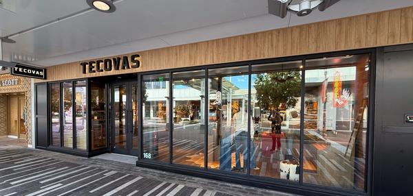 Storefront of Tecovas with large glass windows, wood paneling above, and reflections of nearby shops and street visible outside.