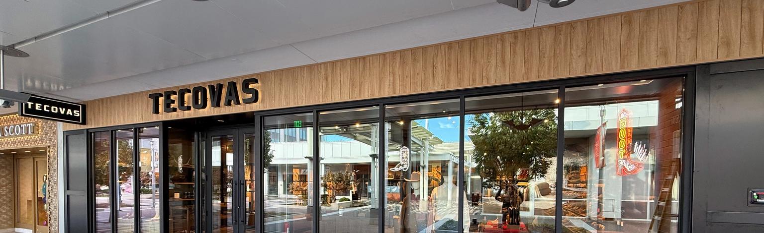 Storefront of Tecovas with large glass windows, wood paneling above, and reflections of nearby shops and street visible outside.