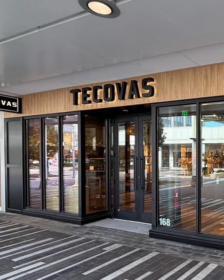 Storefront of Tecovas with large glass windows, wood paneling above, and reflections of nearby shops and street visible outside.
