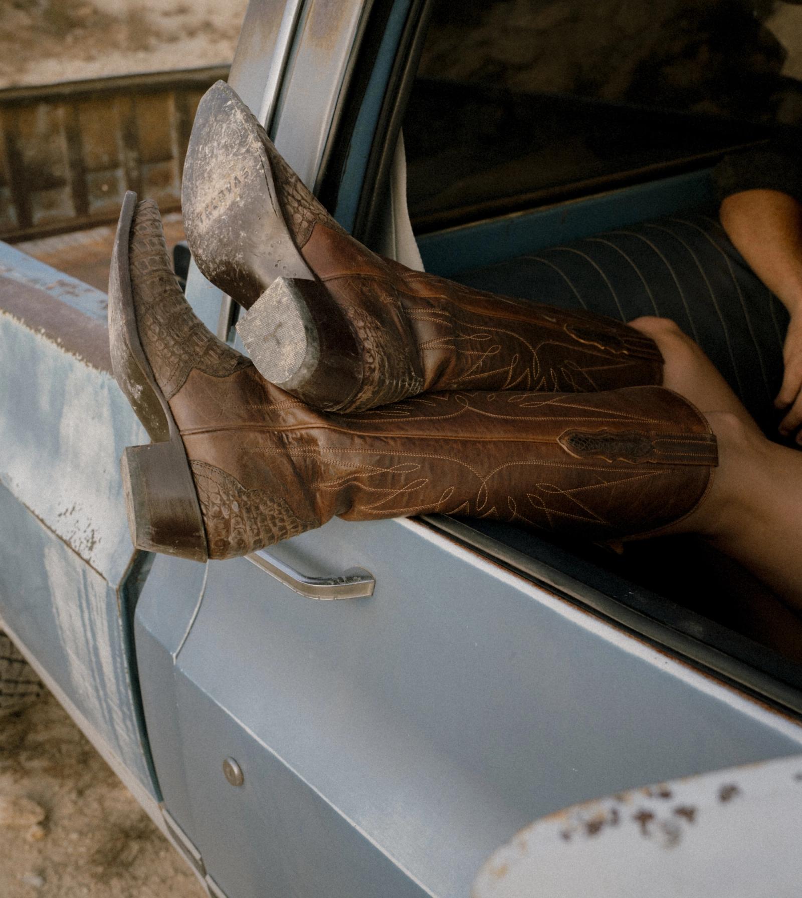 Pair of worn brown Tecovas cowboy boots resting on the open window of a pale blue pickup truck