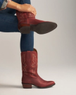 The Britt in Redwood Goat. A person wearing blue jeans and a patterned shirt models a pair of Britt Goat red cowboy boots with decorative stitching, crossing one leg over the other.
