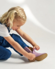 Young child with blonde hair wearing a white shirt and denim overalls zips up a lavender boot while sitting on the floor.