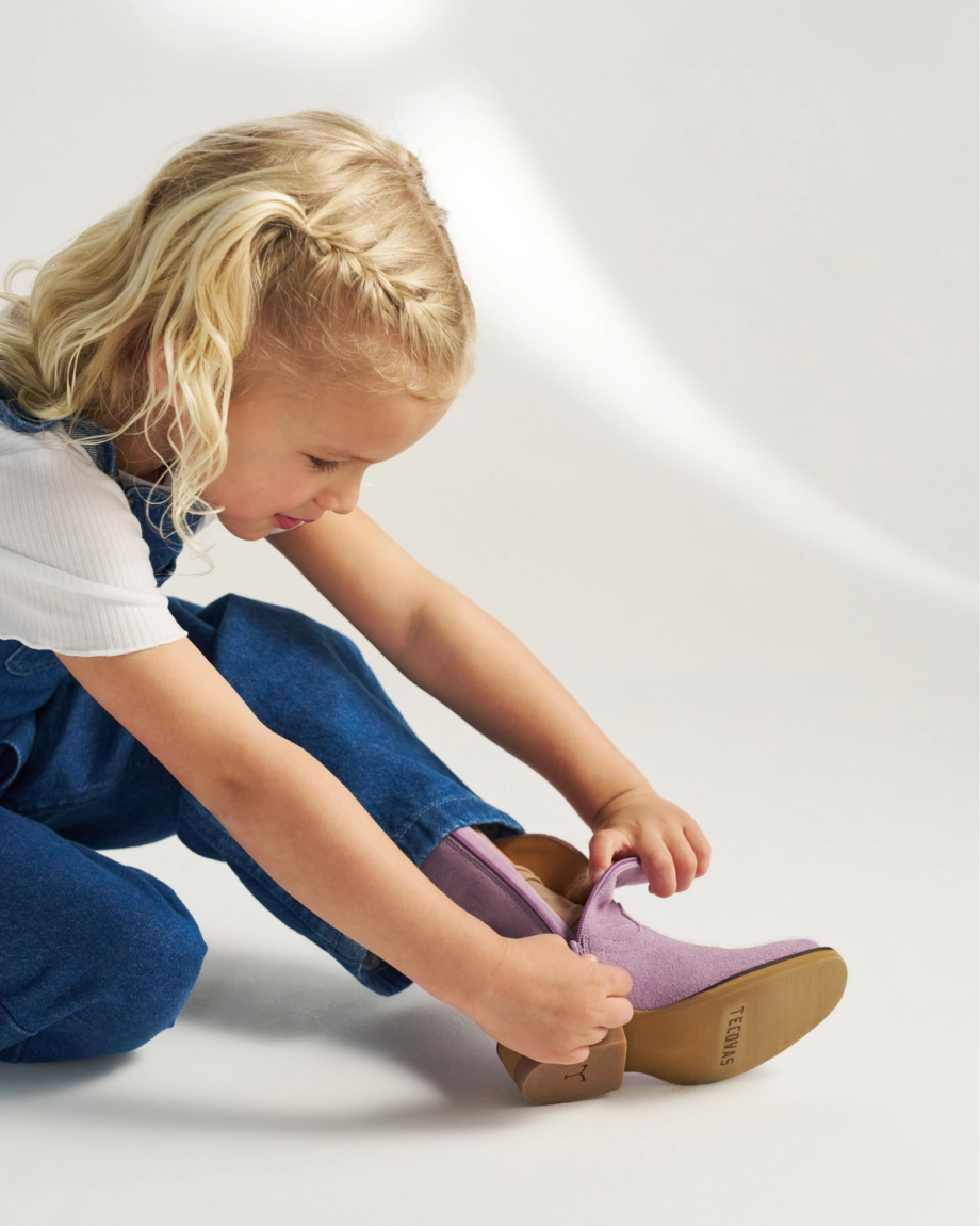Young child with blonde hair wearing a white shirt and denim overalls zips up a lavender boot while sitting on the floor.