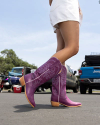 A person wearing purple cowboy boots and white shorts stands on a paved surface with parked vehicles and trees in the background.