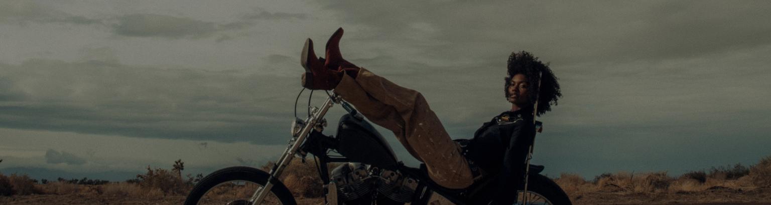 A person reclines on a parked motorcycle in a desert landscape, with legs resting on the handlebars and a cloudy sky in the background.