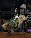 A bull rider wearing protective gear rides a bucking bull in an indoor arena, surrounded by spectators and advertisements.