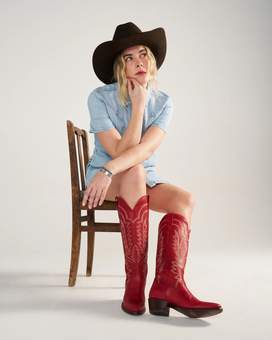 The Abby in Crimson Cowhide. A woman in a denim dress, crimson cowhide boots, and a black cowboy hat sits on a wooden chair, looking thoughtfully to the side.