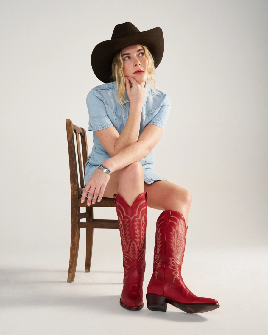 The Abby in Crimson Cowhide. A woman in a denim dress, crimson cowhide boots, and a black cowboy hat sits on a wooden chair, looking thoughtfully to the side.