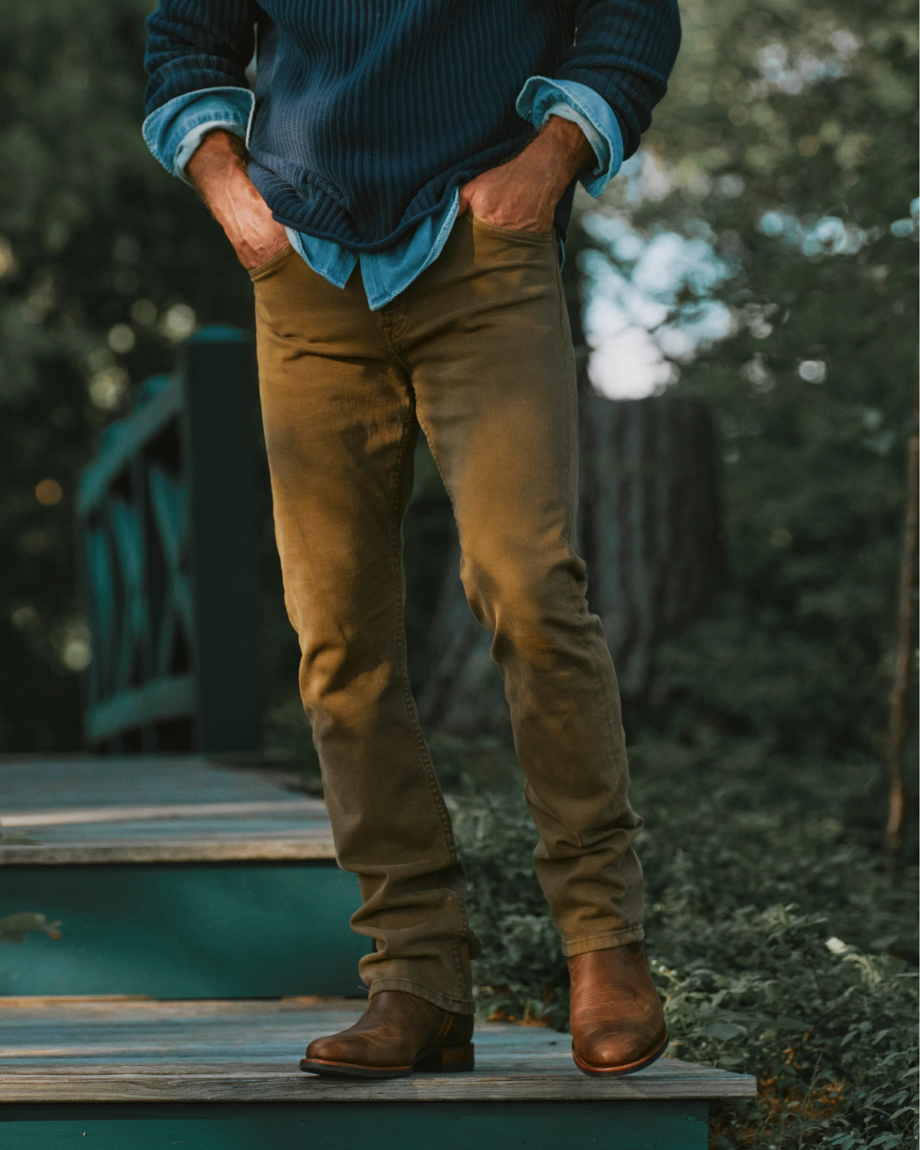 The Wayne in Maplewood Cowhide. A person stands outdoors on steps, wearing brown Maplewood Cowhide pants, a blue sweater layered over a denim shirt, and brown boots. Hands are in their pockets. Their face is not visible.