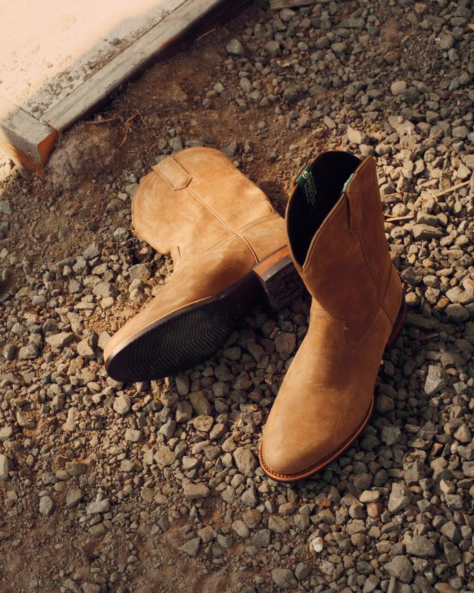 A pair of tan suede boots is placed on gravel near a concrete edge, with sunlight casting shadows on the scene.