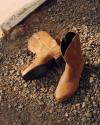 A pair of tan suede boots is placed on gravel near a concrete edge, with sunlight casting shadows on the scene.