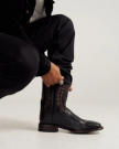 Person wearing black pants and jacket kneeling down, adjusting a black cowboy boot with decorative stitching, against a plain light background.
