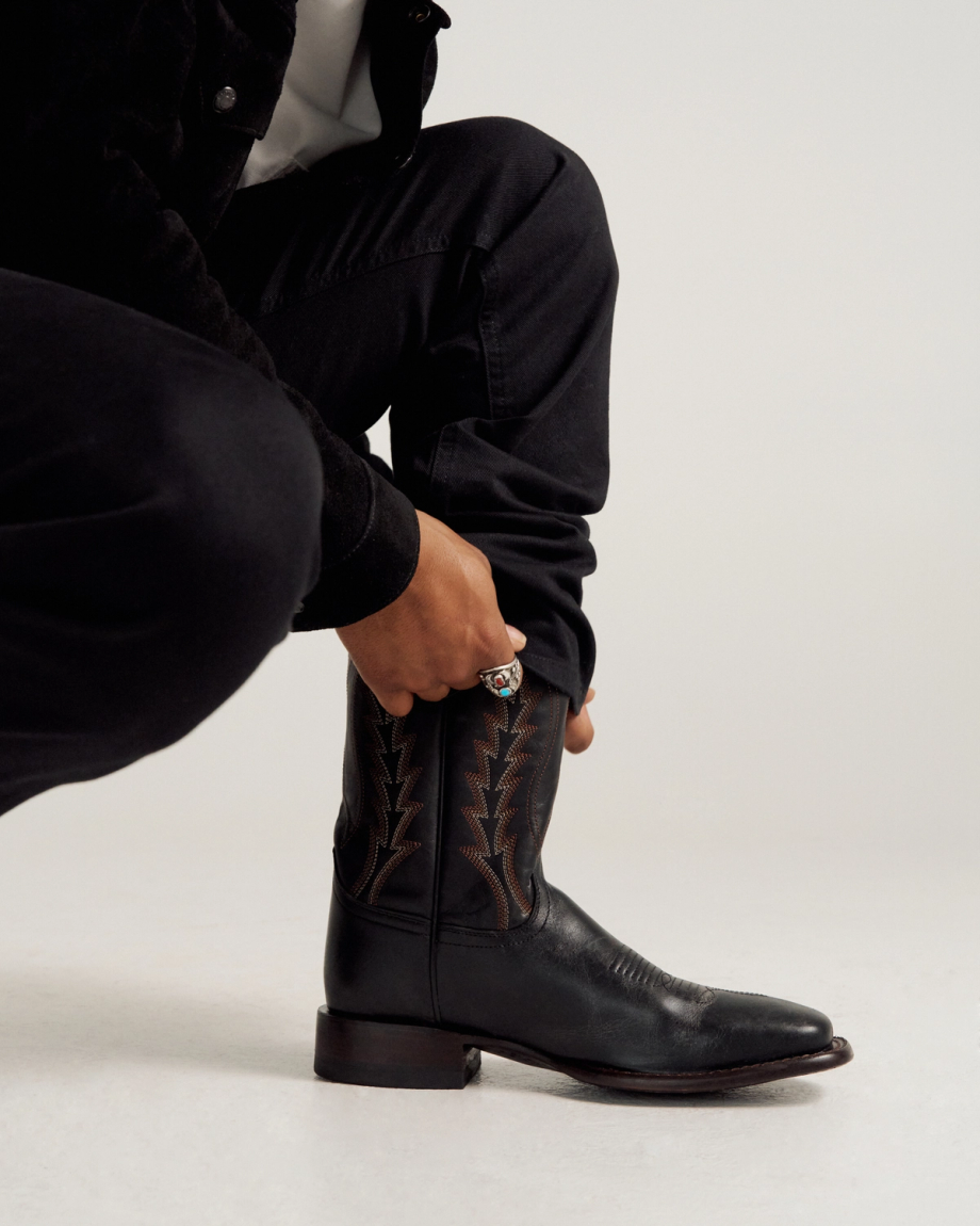 Person wearing black pants and jacket kneeling down, adjusting a black cowboy boot with decorative stitching, against a plain light background.