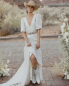 A woman wearing a cowboy hat and white dress stands outdoors on a brick pathway, surrounded by white floral arrangements. She is holding the hem of her dress and wearing white cowboy boots.