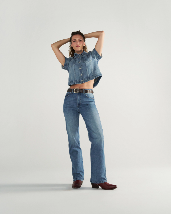 A person stands against a plain background wearing a short-sleeve denim shirt, blue jeans, a black belt, and brown boots, with arms raised and hands behind their head.