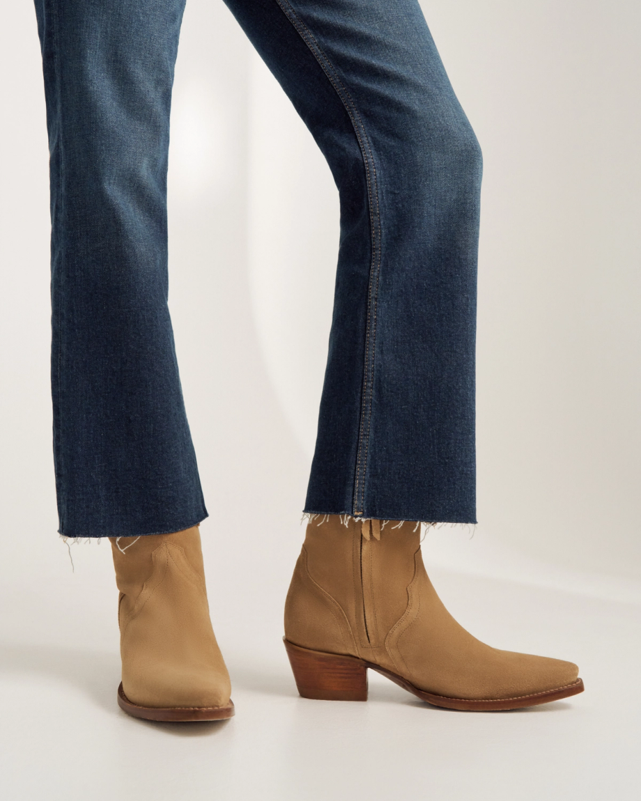 Person wearing blue denim jeans with raw hems and tan heeled western-style ankle boots, standing on a light-colored floor.