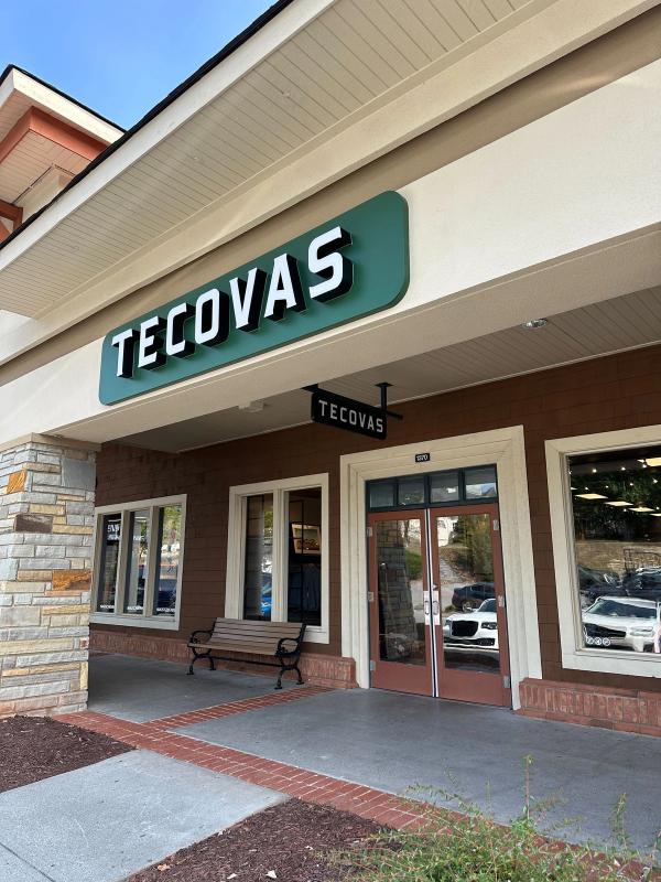Storefront of Tecovas, featuring a green sign with white lettering above the entrance. A bench is placed outside the store, and the glass doors reveal the interior.