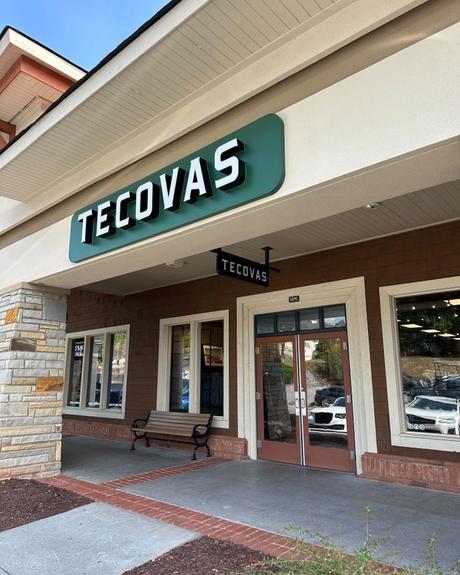 Storefront of Tecovas, featuring a green sign with white lettering above the entrance. A bench is placed outside the store, and the glass doors reveal the interior.