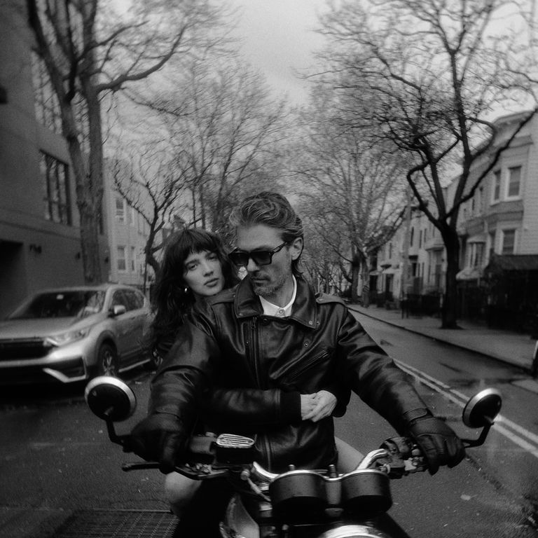 A man and a woman ride a motorcycle down a residential street lined with bare trees and parked cars.