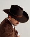 A man wearing a brown cowboy hat and a brown jacket looks slightly downward against a neutral background.