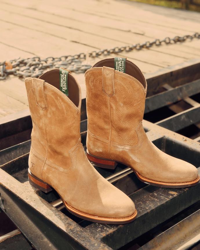 A pair of tan suede cowboy boots with green tags, resting on a metal grate outdoors.