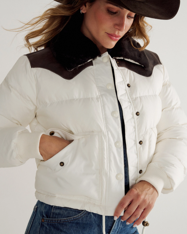 A person wearing a white puffer jacket with black collar detailing, blue jeans, and a black wide-brimmed hat, with one hand in their pocket.