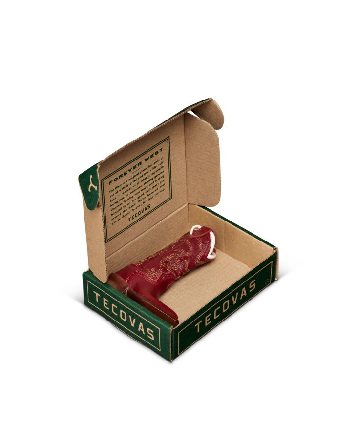 A small red cowboy boot displayed inside an open Tecovas-branded cardboard box with printed text on the lid.