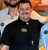 Three people standing together, two partially visible. Central figure wearing a black shirt with a name badge and bolo tie. All are smiling and appear to be in a casual indoor setting.