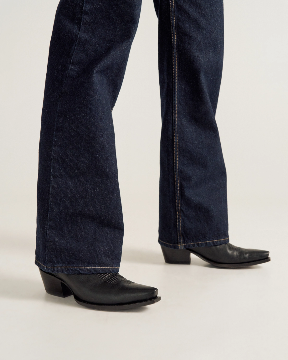 A person wearing dark blue bootcut jeans and black heeled boots stands on a light-colored floor.